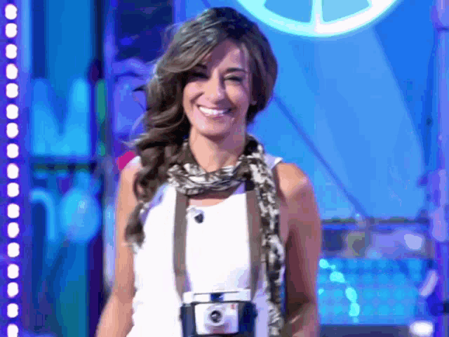 a woman wearing a white tank top and a scarf smiles while holding a camera