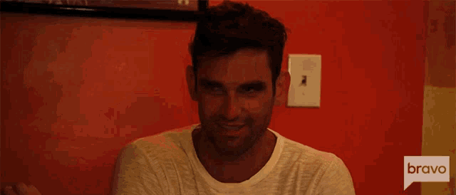 a man sitting in front of a red wall with a bravo logo on the bottom right