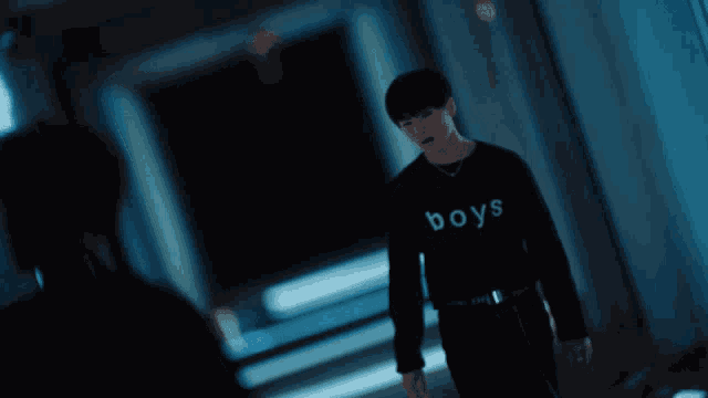 a man wearing a black shirt that says boys stands in a dark hallway