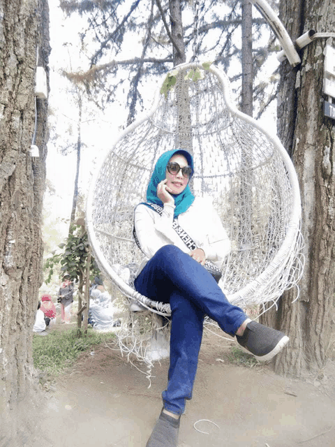a woman wearing a hijab and sunglasses is sitting in a hanging chair