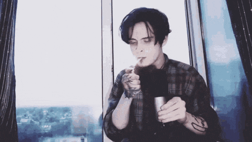 a man is smoking a cigarette in front of a window while holding a cup of coffee .