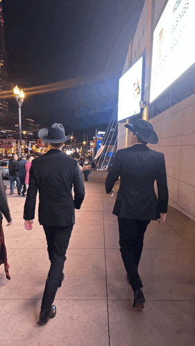 two men in black suits and hats walk down a sidewalk