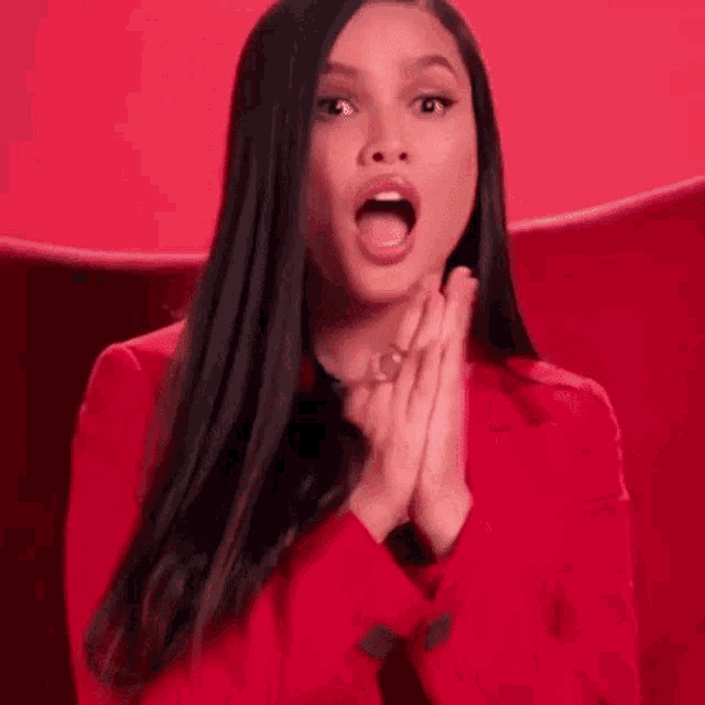 a woman in a red jacket is sitting in a chair with her mouth open and her hands together .