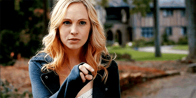 a woman in a blue jacket is standing in front of a house and looking at the camera .