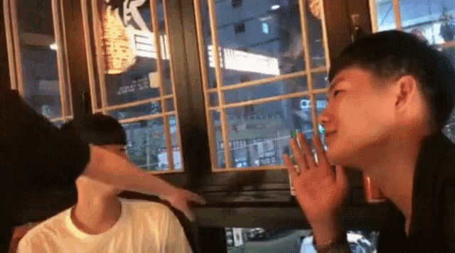 two men are sitting at a table with their hands on their faces in front of a window
