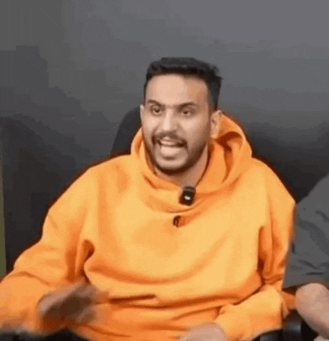 a man with a beard wearing an orange hoodie is sitting in a chair and making a funny face .