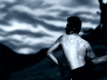 a shirtless man with a tattoo on his arm stands in front of a cloudy sky