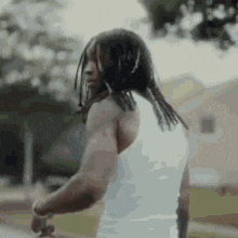 a man with dreadlocks is wearing a white tank top and is standing in front of a house .