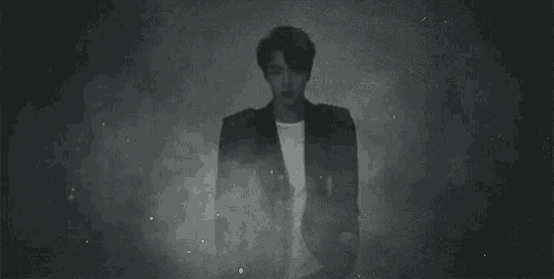 a black and white photo of a man in a suit standing in a dark room surrounded by smoke .
