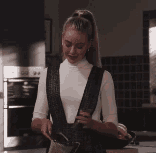 a woman wearing a white turtleneck and black overalls is standing in a kitchen
