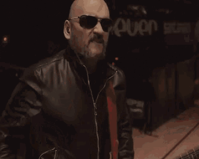 a bald man wearing sunglasses and a leather jacket is playing a guitar