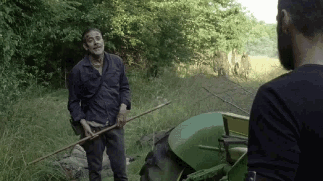 a man in a blue shirt is standing next to a green tractor holding a stick
