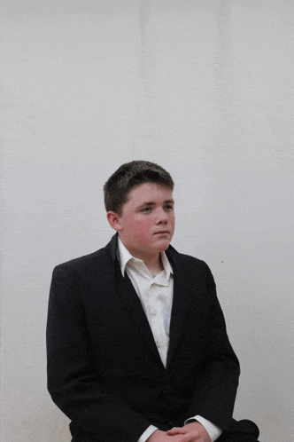 a young man in a black suit and white shirt sits with his hands folded