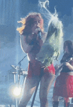 a woman with red hair is singing into a microphone while another woman with green hair is dancing