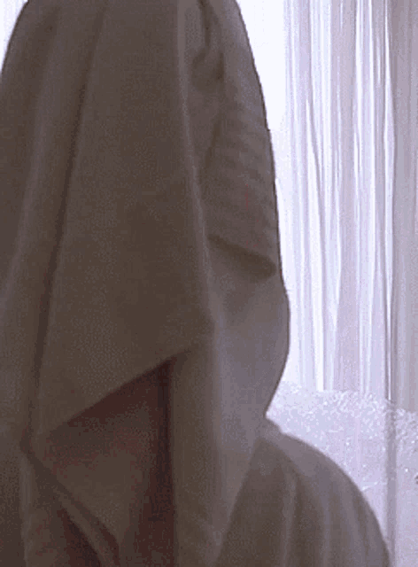 a person with a towel draped over their head looks out a window