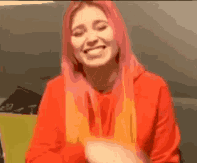 a woman with pink hair is wearing a red hoodie and smiling while holding a carrot .