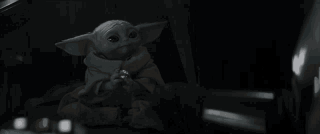 a baby yoda from star wars is sitting in a dark room holding a sword .