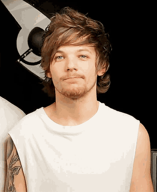 a man with a beard and a white tank top looks at the camera