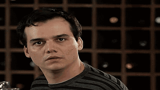 a man in a striped shirt is looking at the camera in front of a wine rack