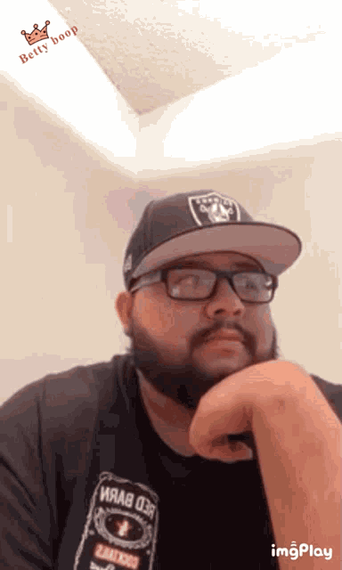 a man with a beard wearing a raiders hat and glasses looks at the camera