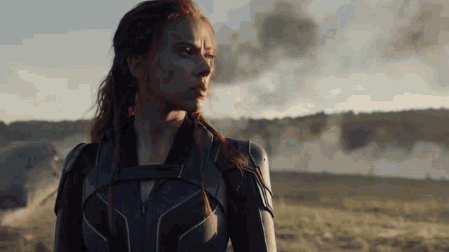 a woman in a black suit stands in a field with smoke in the background