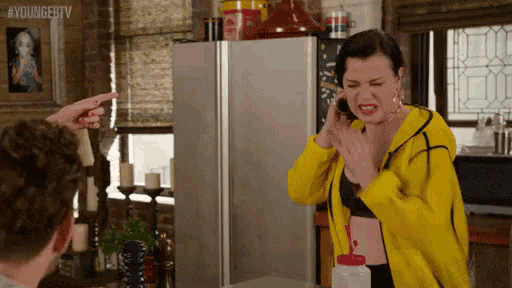 a woman in a yellow jacket is talking on a cell phone while standing in front of a refrigerator .