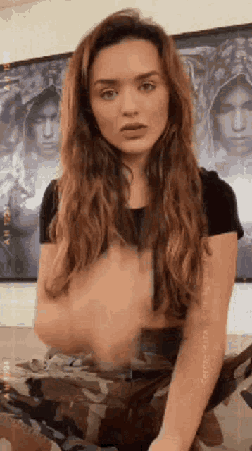 a woman in a black shirt and camouflage pants is sitting on a couch in front of a painting .