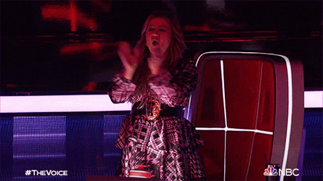 a woman in a plaid dress applauds while sitting in front of a nbc chair