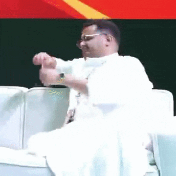 a man wearing glasses and a white robe is sitting on a couch and looking at his watch .