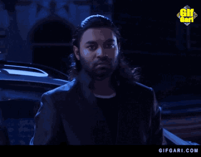 a man with long hair and a beard is standing in front of a car at night ..