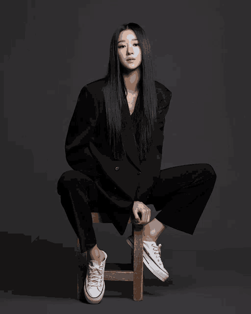a woman in a suit sits on a wooden stool with her legs crossed