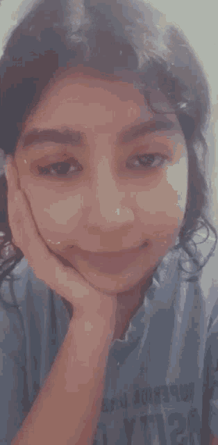 a young girl is smiling with her hand on her chin . she is wearing a blue shirt that says lyt128 on it .