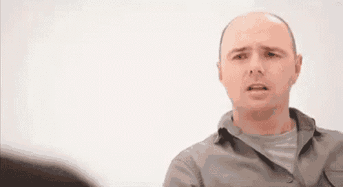 a bald man in a grey shirt is making a funny face while talking to another man .