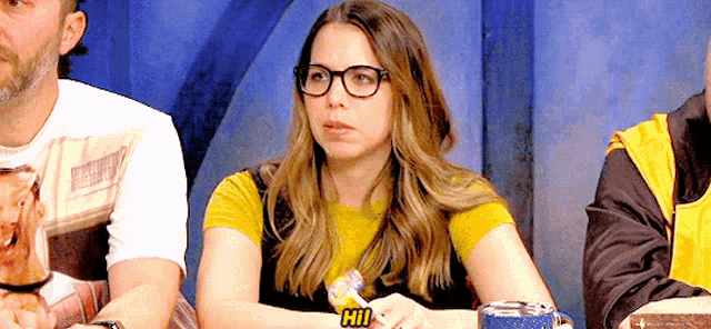 a woman wearing glasses and a yellow shirt is sitting at a table with a lollipop in her hand .