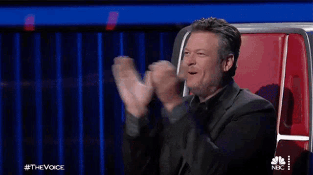 a man in a suit is sitting in a red chair and clapping his hands .