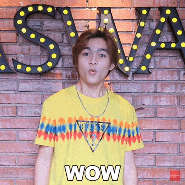 a man wearing a yellow guess shirt is making a surprised face in front of a brick wall