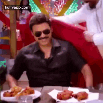 a man wearing sunglasses and a mustache is sitting at a table with a plate of food .