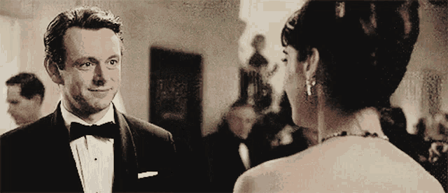 a man in a tuxedo and bow tie looks at a woman in a white dress