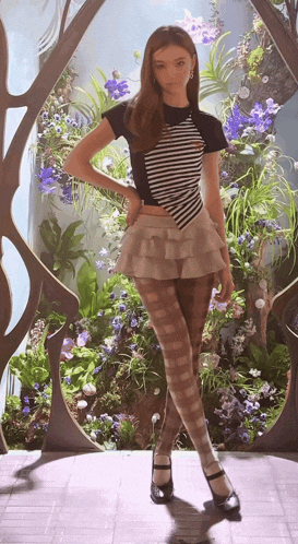 a woman in a striped shirt and ruffled skirt stands in front of a display of flowers