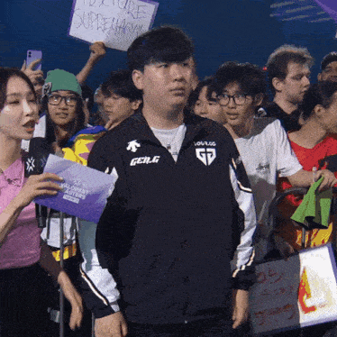 a man wearing a gen.g jacket stands in a crowd of people