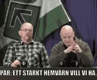 two men are sitting at a table in front of a flag .