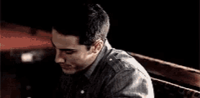 a man in a gray shirt is sitting in a dark room with his head on another man 's shoulder .