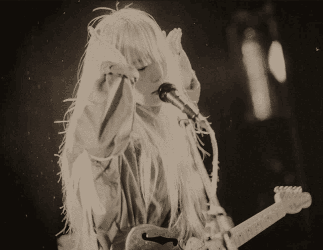 a woman with long white hair is singing into a microphone while playing a guitar