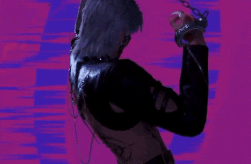 a man in a black jacket and chains is dancing in front of a pink and blue background .