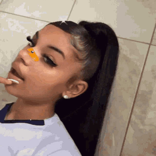 a girl with long black hair and a ponytail is taking a selfie with a yellow sticker on her nose .