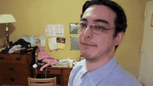 a man wearing glasses stands in front of a messy room