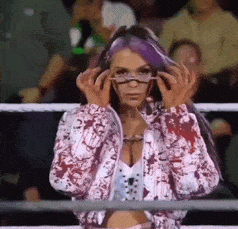 a woman with purple hair is standing in a wrestling ring wearing glasses .
