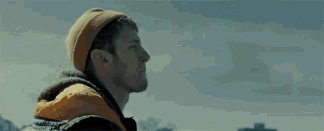 a man wearing an orange jacket and a yellow beanie is standing in front of a mountain .