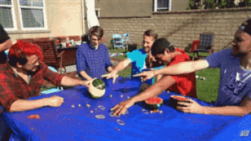 a group of people are sitting around a table with watermelons