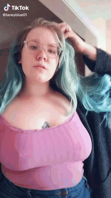 a woman with blue hair and glasses is wearing a pink top and jeans .